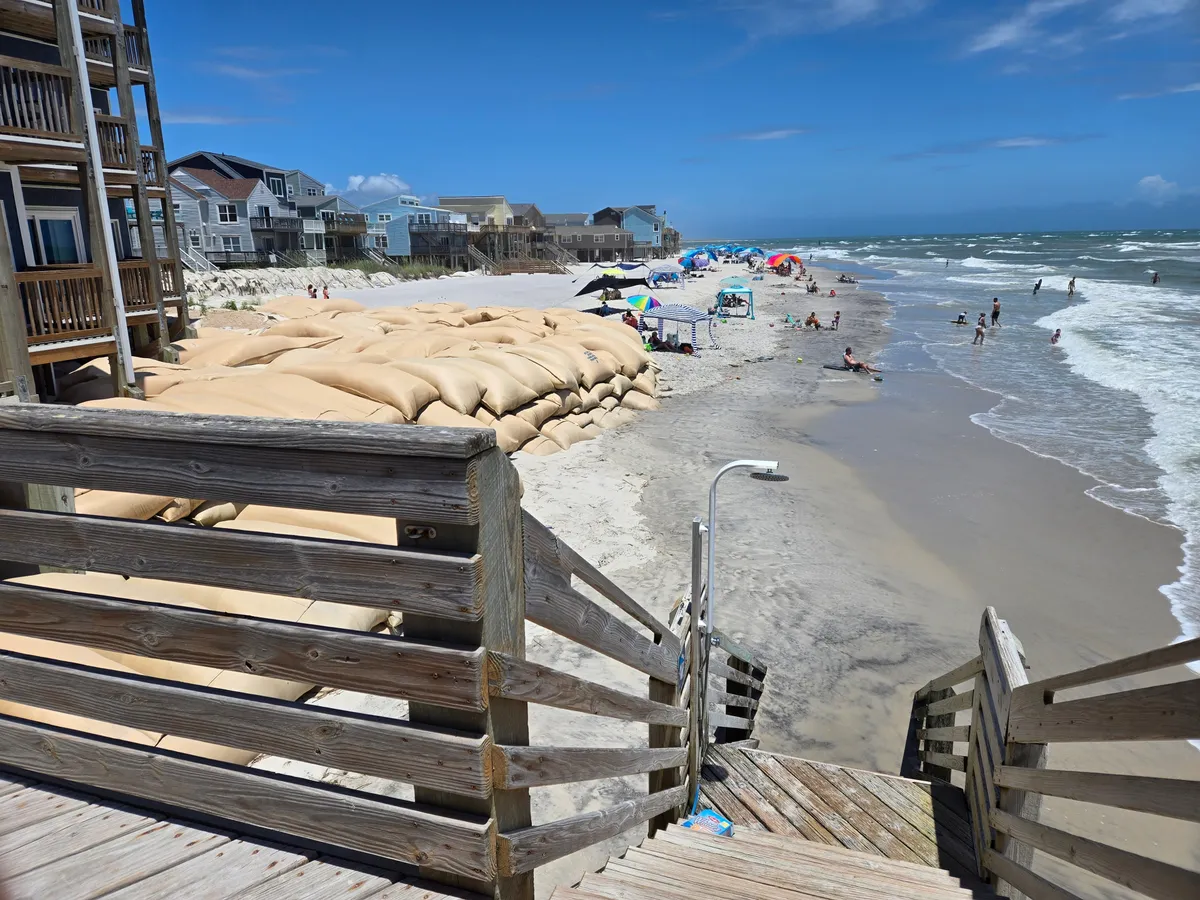 A vibrant beachfront scene with families and groups enjoying the sand and surf. Colorful umbrellas and beach gear dot the sand, with people sunbathing, swimming, playing games, and building sandcastles. The beach houses in the background reinforce the proximity to accommodation and easy access to seaside activities.