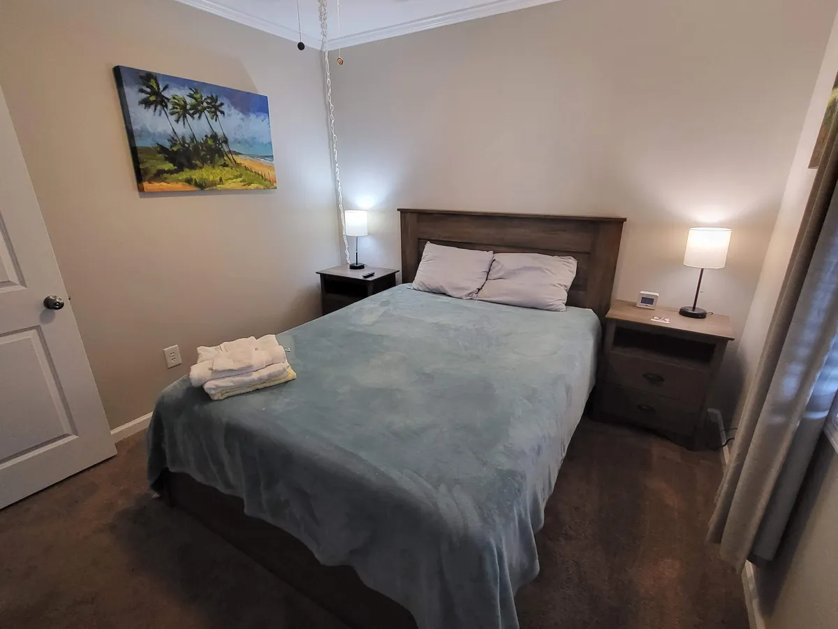 A calm and inviting bedroom scene with a well-made bed, nightstands with lamps, and a piece of coastal-inspired artwork above the headboard. The room glows with natural morning light, creating a soothing ambiance.