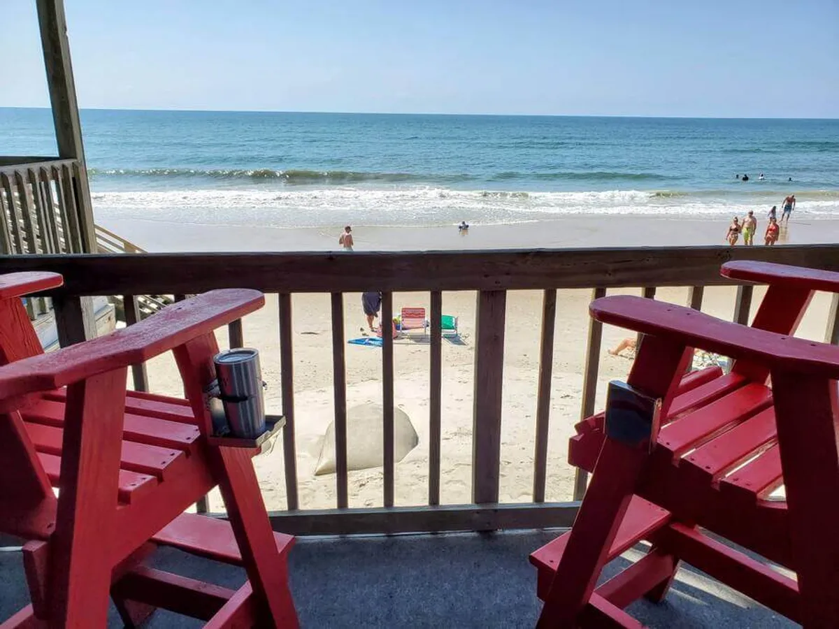 The image showcases a pair of bright red Adirondack chairs on a private balcony, overlooking endless stretches of sand and the sparkling Atlantic Ocean. Guests are seen relaxing in the chairs, enjoying drinks, while children play in the sand and people swim and sunbathe along the pristine North Topsail Beach shoreline.