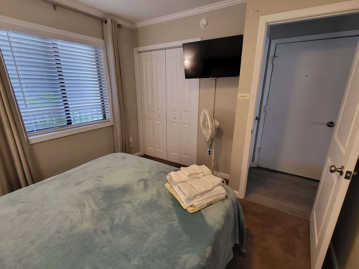 A serene bedroom scene with a soft blue bedspread, stacked towels, and a window that allows light to stream in. The atmosphere is calm and cozy, suggesting a relaxing retreat ideal for unwinding after a day at the beach.