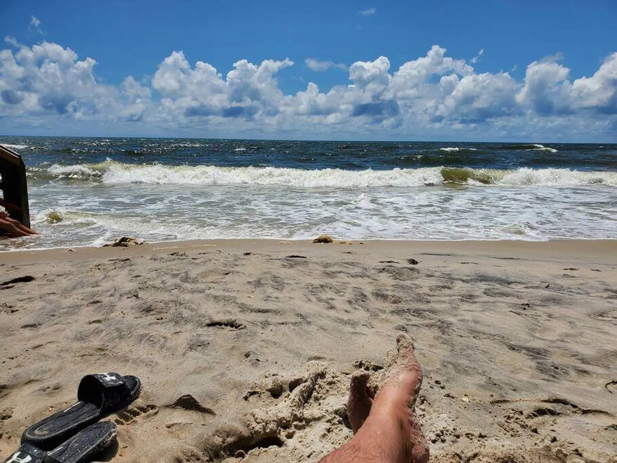 A close-up of sandy beach with gentle ocean waves rolling in, clouds painted across the horizon, and a single foot in the foreground. The scene conveys active engagement with the shoreline, from walking to swimming and collecting seashells.