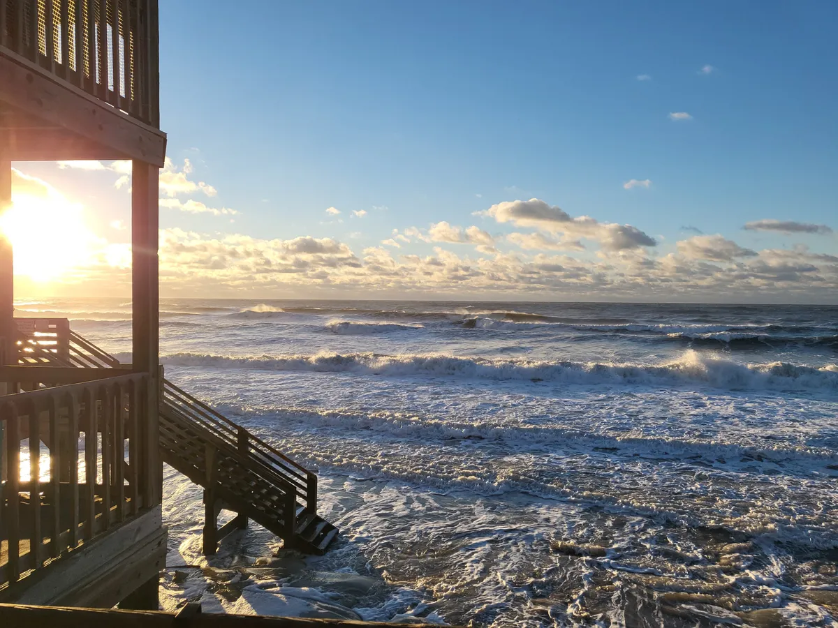A dramatic sunrise unfolds over the ocean, with vibrant colors reflecting on the gentle waves. A wooden staircase leads down to the sand, hinting at the possibility of beach walks and intimate moments just outside the balcony. The perspective emphasizes proximity to the shoreline and the beauty of natural light at different times of day.