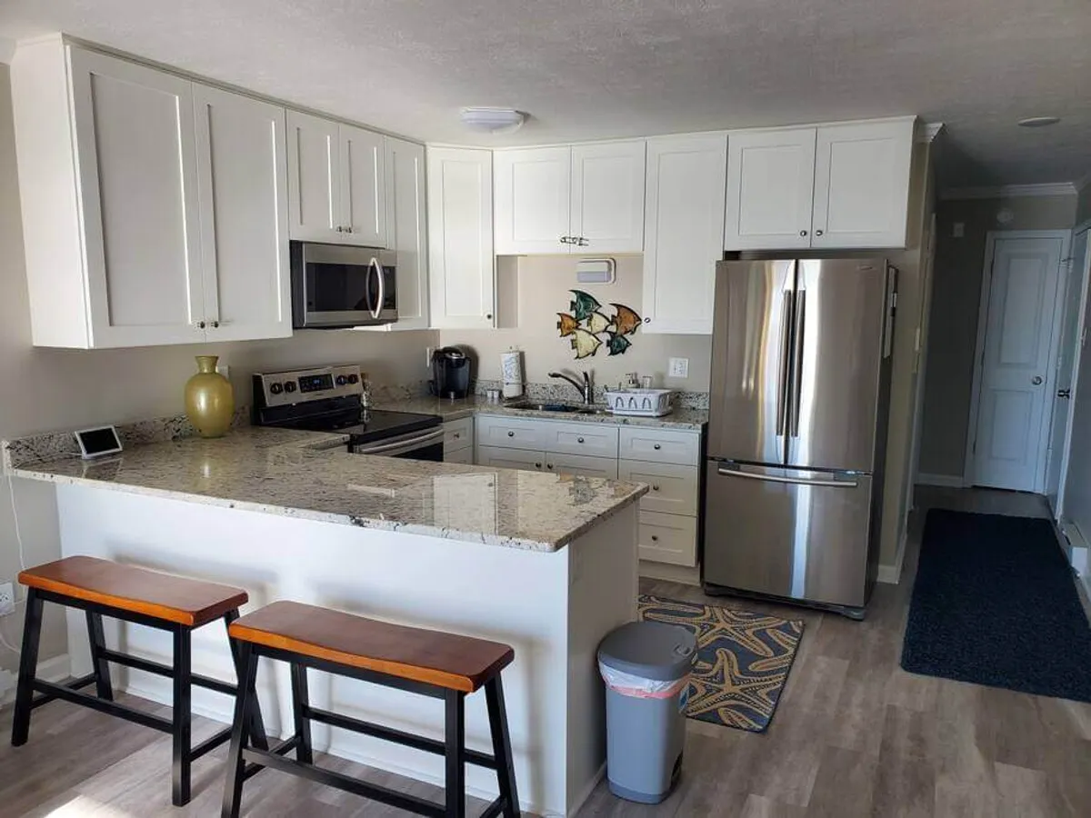 A bright, welcoming kitchen with a large granite countertop, sleek stainless steel appliances, and vibrant fish artwork on the wall. Bar stools are positioned at the counter, inviting casual conversation or a quick meal. Sunlight fills the space, highlighting a design meant for both cooking and gathering.