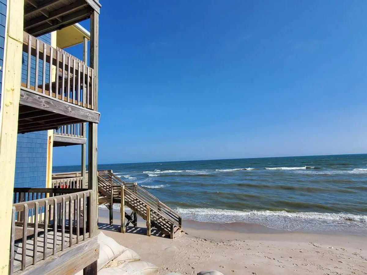 This view looks out from a beachfront deck or balcony, bordered by wooden stairs leading down to the shore. The waves roll in nearby, and the vantage point offers a sweeping perspective of the ocean and sand, inviting the viewer to imagine stepping directly onto the beach from the retreat.