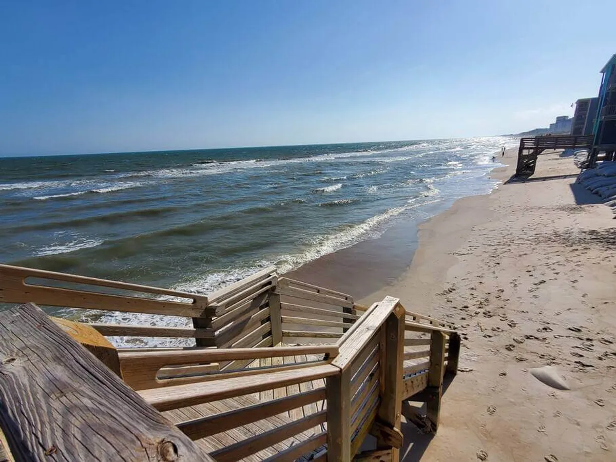 A set of wooden stairs leads down to a gently curving beach where waves lap at the shore, and distant beach houses dot the horizon. The scene feels inviting and intimate, conjuring images of couples taking strolls along the sand or enjoying the sunset together.