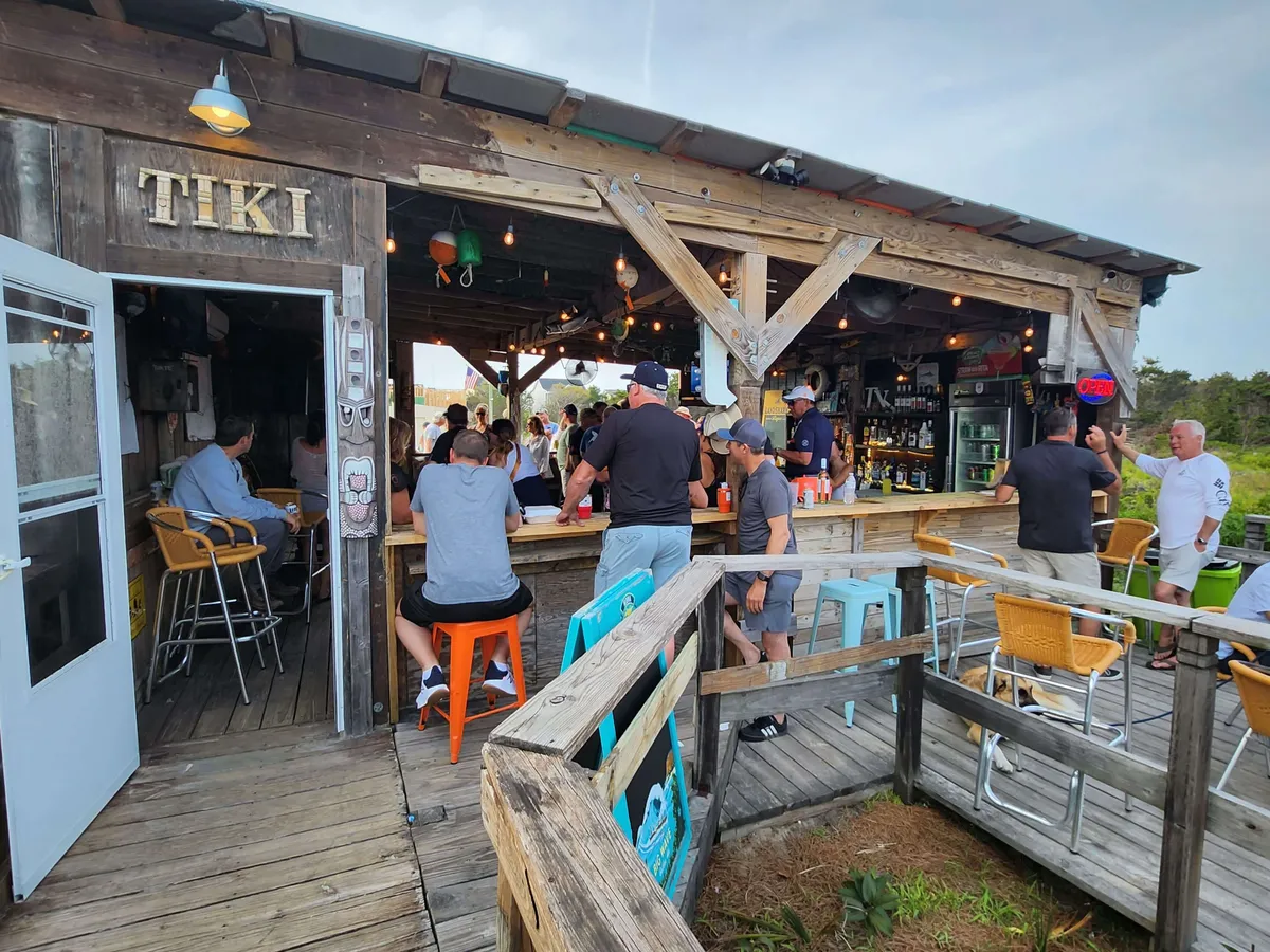 A festive tiki bar deck overlooks the water, with colorful chairs, a tiki bar sign, and people socializing at sunset. The scene is lively yet relaxed, capturing the essence of a fun, memorable date night by the ocean.