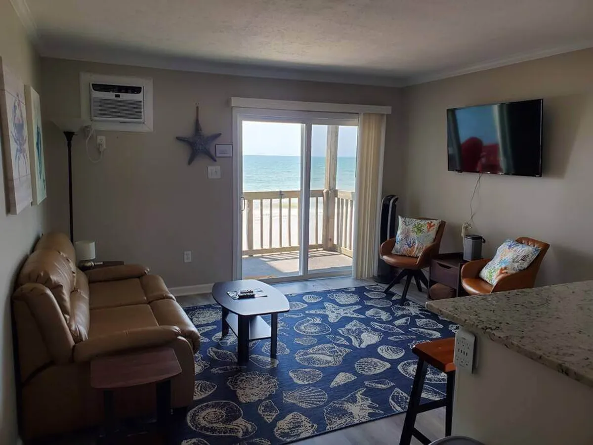 A cozy living area featuring sliding glass doors that open directly onto the beach, comfortable seating with colorful cushions, a decorative starfish on the wall, and a bright, airy atmosphere. The ocean view becomes an integral part of the space, and the room feels inviting for relaxing, socializing, or enjoying entertainment.