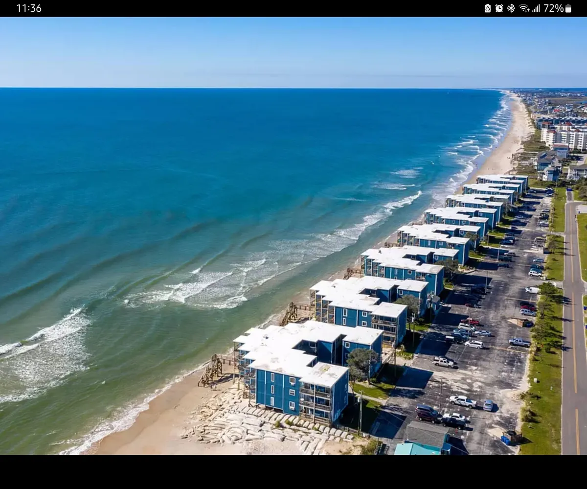 This image captures a tranquil view of ocean waves rolling onto the sandy beach, with beachfront buildings and a parking lot in the background. The setting evokes the peaceful ambiance of a coastal retreat, ideal for relaxation and romantic moments. The open expanse of water under soft lighting suggests both sunrise and sunset possibilities, with easy access to the shoreline.
