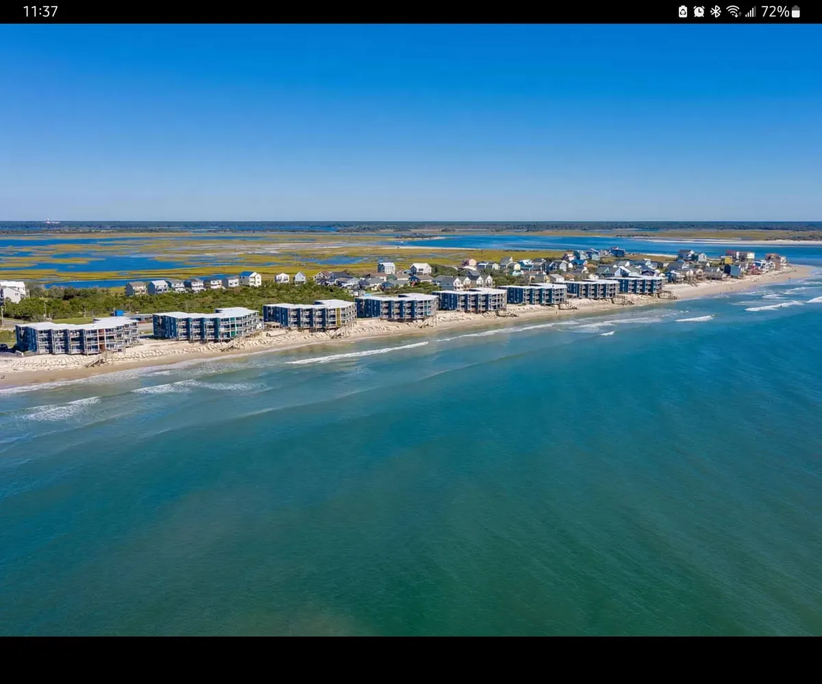 A sweeping view of the coastline featuring beachfront properties overlooking gentle waves and a distant marshland. The expansive blue ocean and clear sky create a serene backdrop, while the scene suggests swimming, sunbathing, and exploring the shore are steps away.