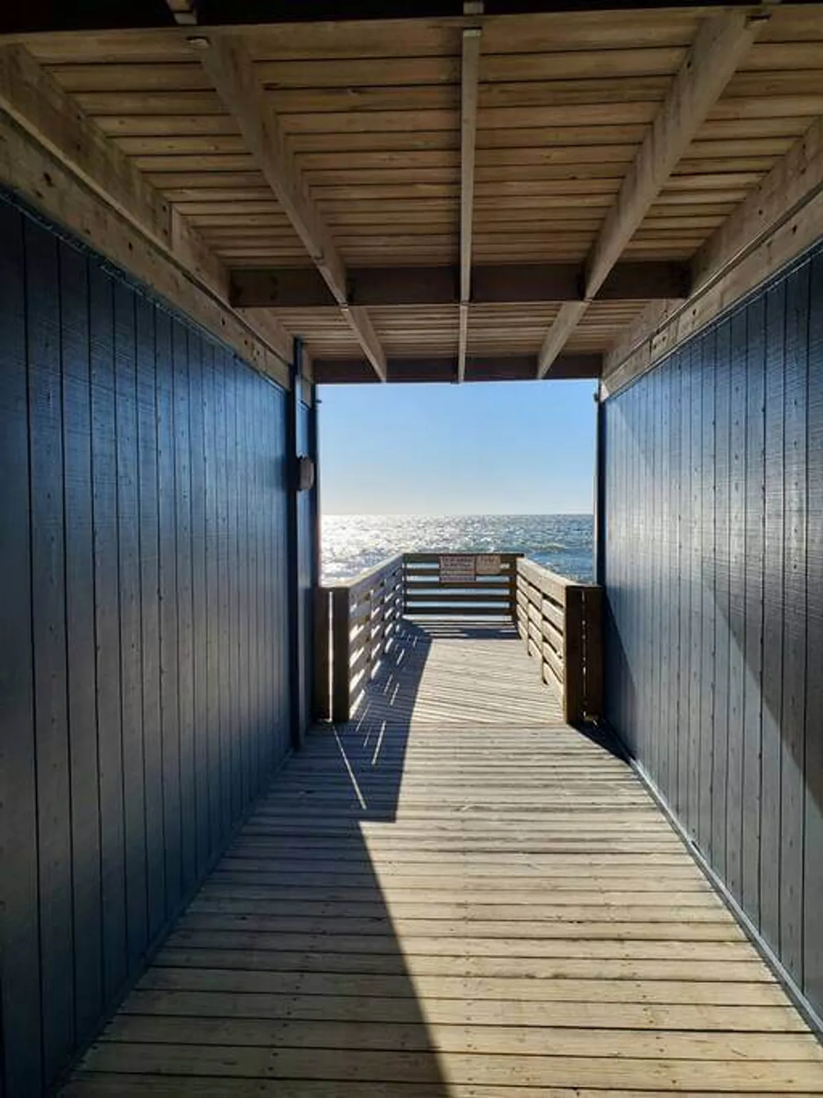 A wooden boardwalk leads directly toward the ocean, bordered by blue walls and opening onto sweeping water views. People are seen walking and relaxing, evoking the sense of easy access to the shore and opportunities for activities like fishing or enjoying the sunrise.
