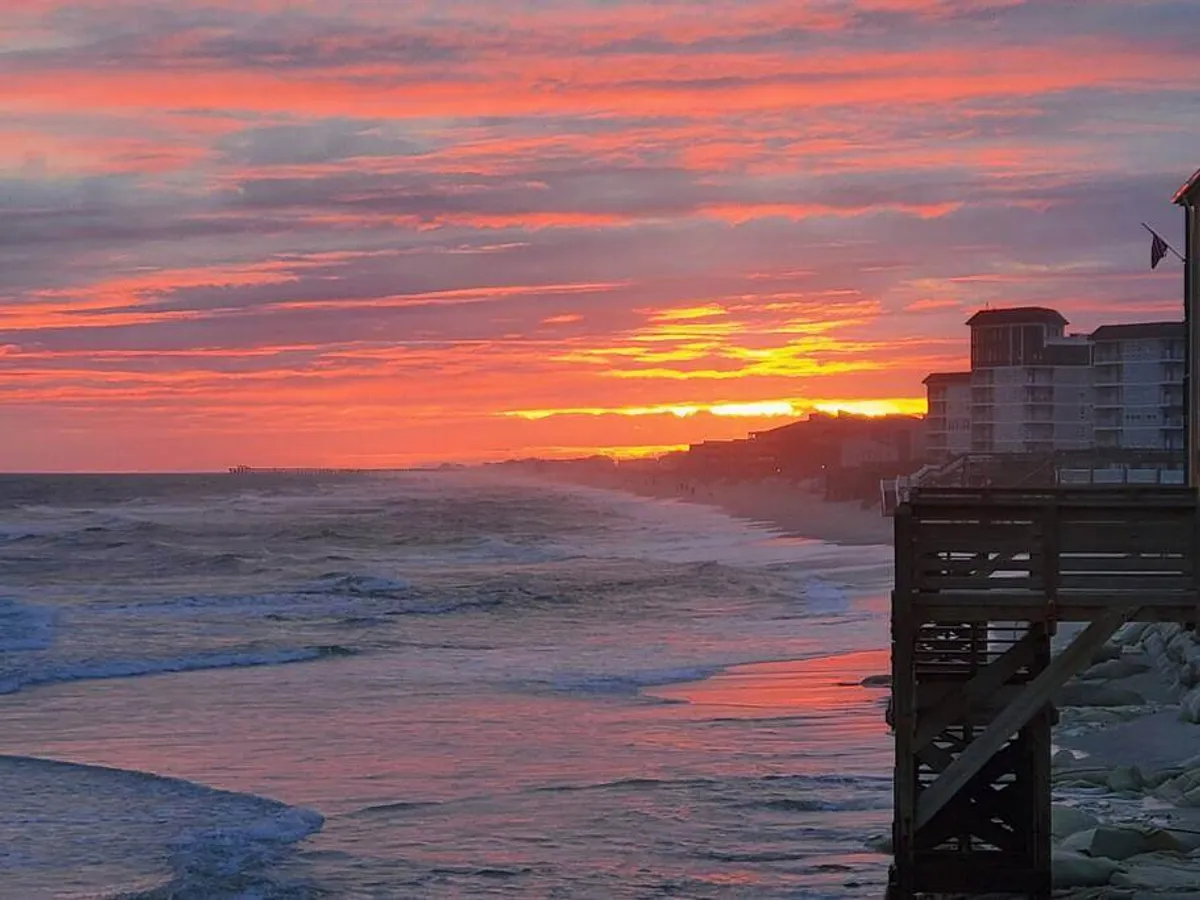 A golden-hued sunset over ocean waves, with a pier stretching out to sea and beachfront buildings silhouetted against the sky. The tranquil mood invites leisurely sunset strolls and peaceful moments by the water, capturing the magic of evening at the beach.